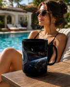 A woman wearing sunglasses is sitting by a pool, with a black mug featuring an image of the Earth and the International Space Station on it.