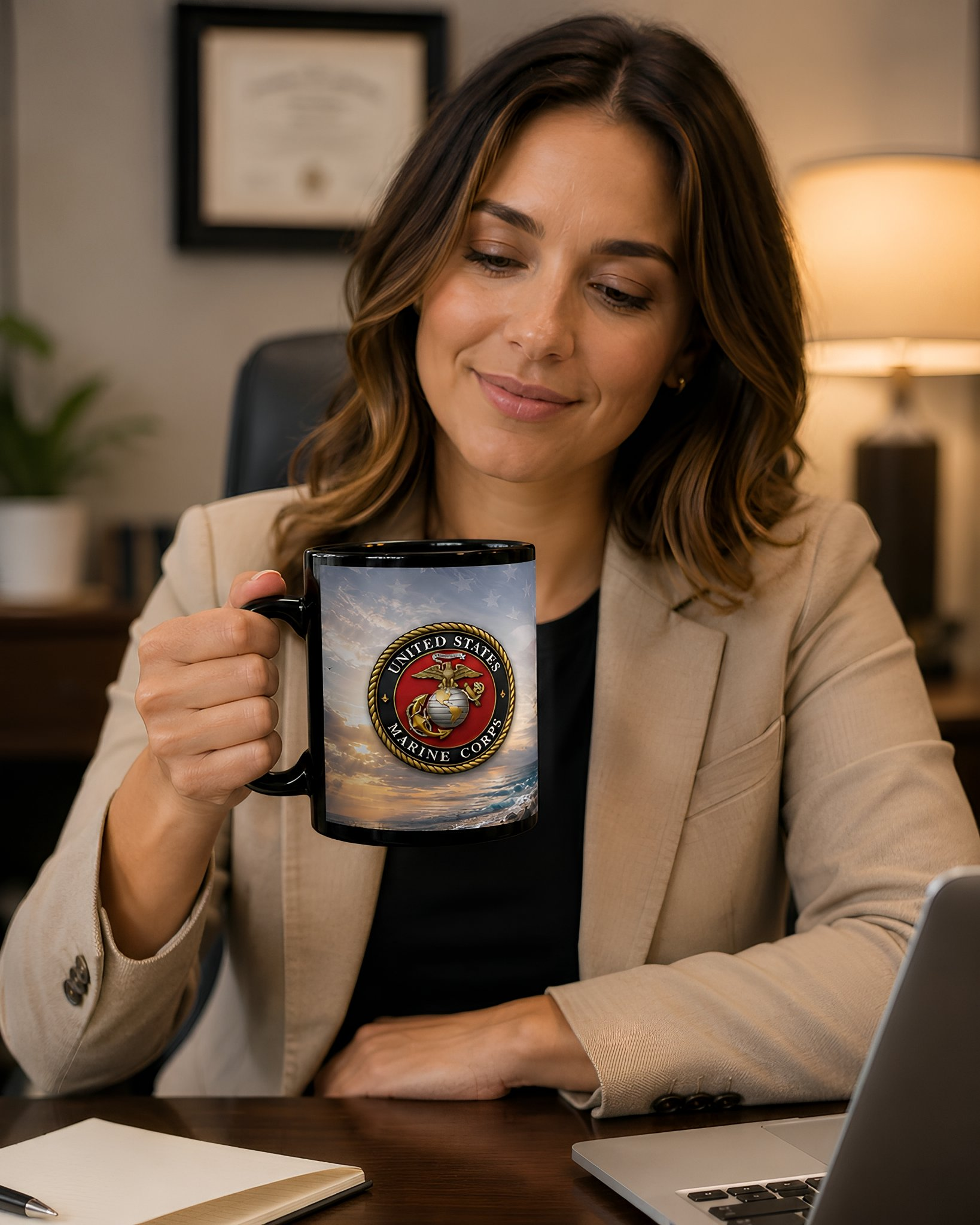 A woman in a beige jacket is sitting at a desk, holding a black mug with a design featuring the United States Marine Corps logo.