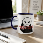 A mug with a cartoon character and text on it, placed on a wooden desk next to a laptop and a notebook.