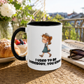 A white mug with a cartoon image of a girl and the text "I used to be somebody, you know" is placed on a table with a plate of pastries, a newspaper, and sunglasses.
