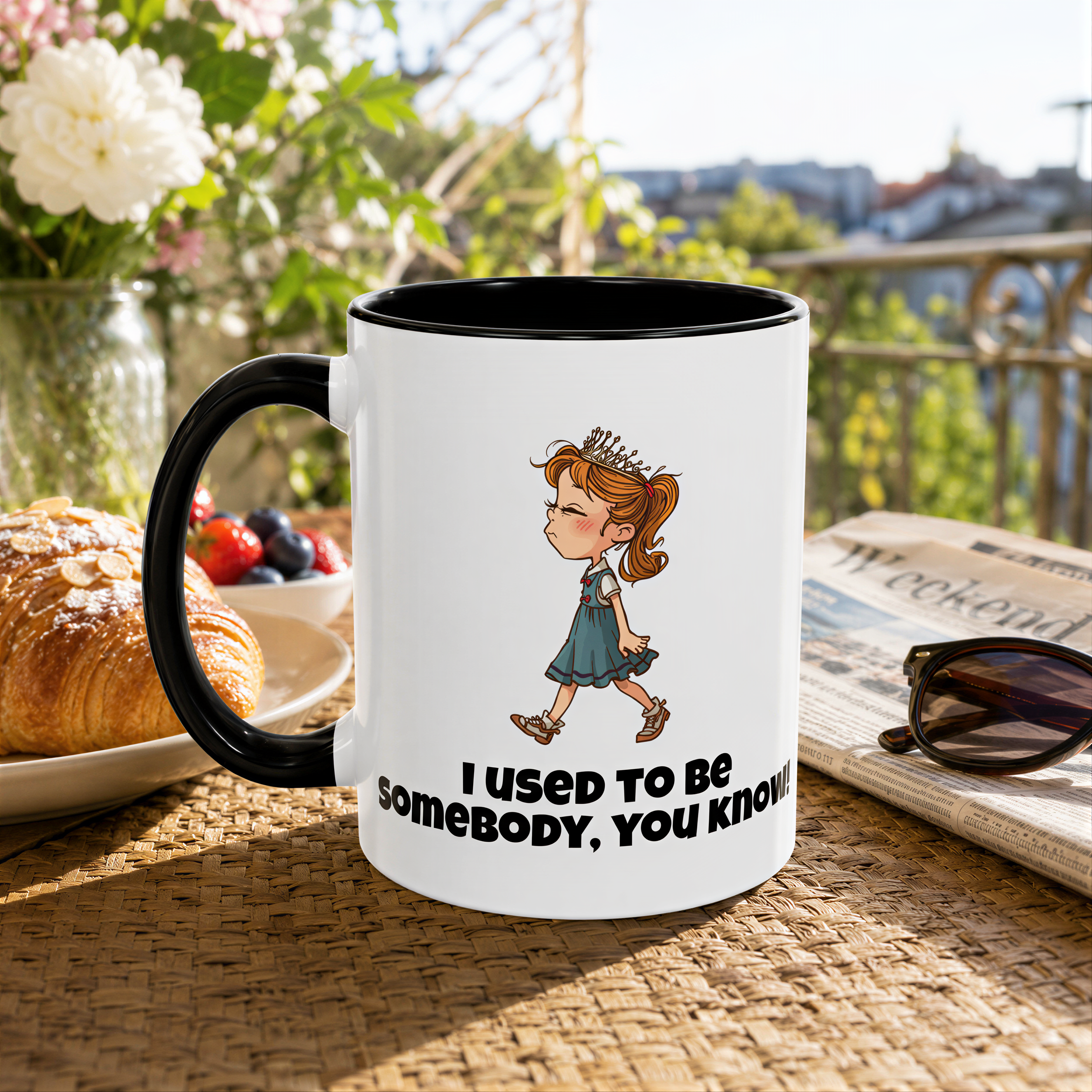 A white mug with a cartoon image of a girl and the text "I used to be somebody, you know" is placed on a table with a plate of pastries, a newspaper, and sunglasses.