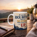 A white mug with a motivational quote sits on a wooden table, with a notebook and pen nearby, and a scenic mountain landscape visible in the background.