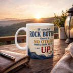 A white mug with a motivational quote sits on a wooden table, with a notebook and pen nearby, and a scenic mountain landscape visible in the background.