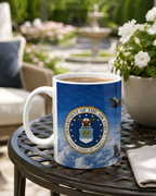A mug with the United States Air Force seal sits on a metal table, with a potted plant and a fountain visible in the background.