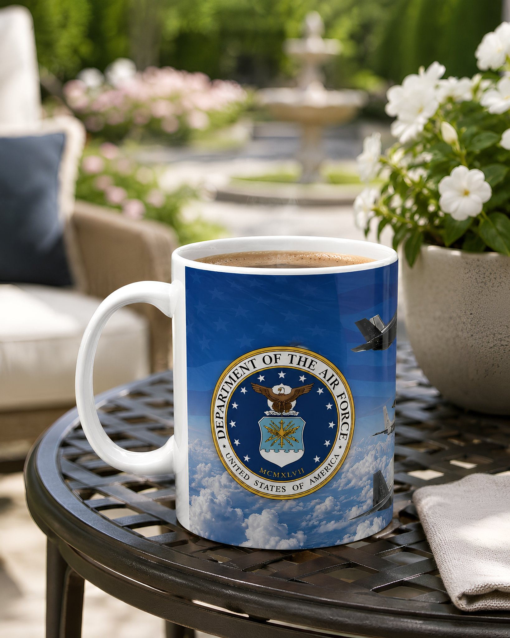 A mug with the United States Air Force seal sits on a metal table, with a potted plant and a fountain visible in the background.