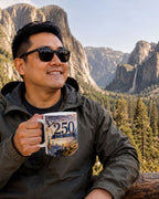 A man in a green jacket is holding a mug with a printed design, smiling and looking towards the camera, with a mountainous landscape in the background.