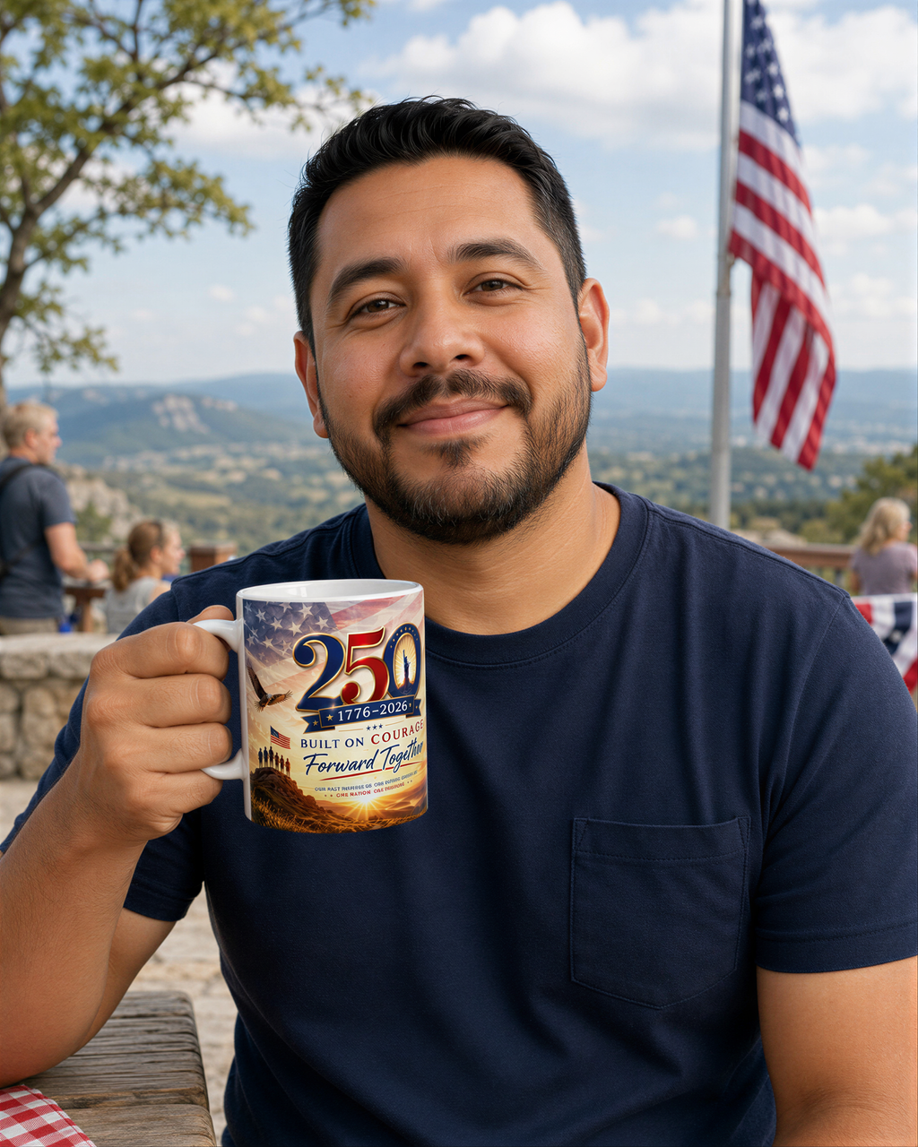 A man is sitting outdoors, holding a mug with a design that says "250" and "1776-2026" on it. He is smiling and looking at the camera. In the background, there is an American flag and a scenic view of mountains.