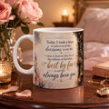 A white ceramic mug with a floral design and text, placed on a wooden table with a vase of pink roses and a lit candle in the background.