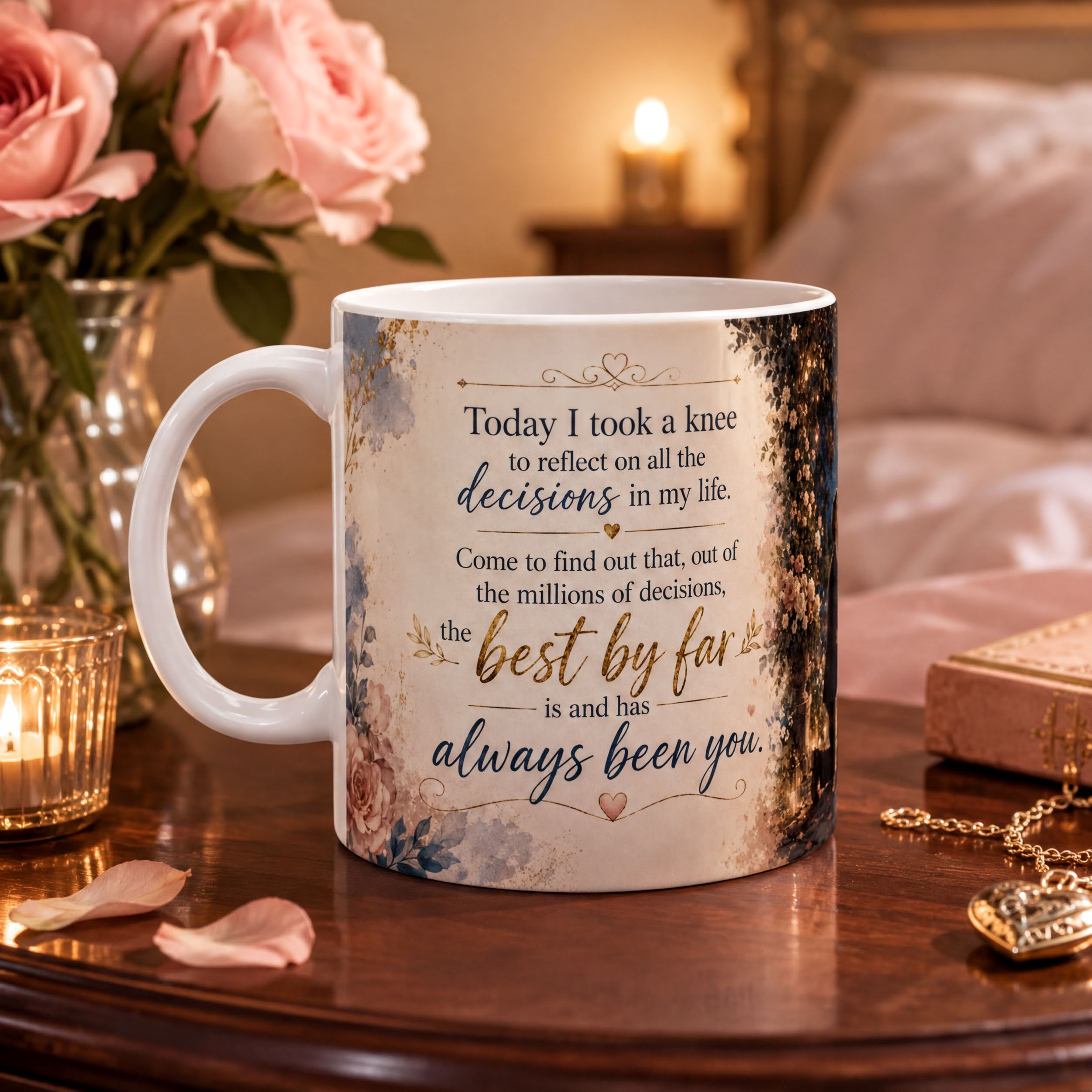 A white ceramic mug with a floral design and text, placed on a wooden table with a vase of pink roses and a lit candle in the background.
