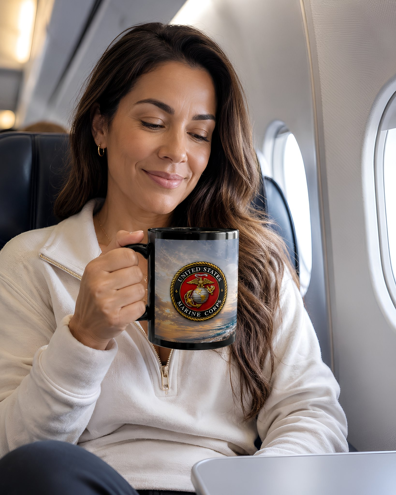 A woman sitting on an airplane, holding a mug with a United States Marine logo, smiling and looking down.