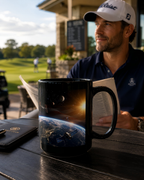 A man in a golf cap and navy polo shirt is sitting at a table with a mug featuring a space scene, reading a book, and looking out at a golf course.