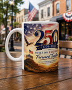 A white ceramic mug with a patriotic design featuring an American flag, eagle, and people standing together, along with the text "25th" and "1776-2026" indicating a 25th anniversary.