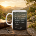 A white mug with a black design featuring a Bible verse and a cross sits on a wooden surface, with a mountain landscape and a sunset in the background.
