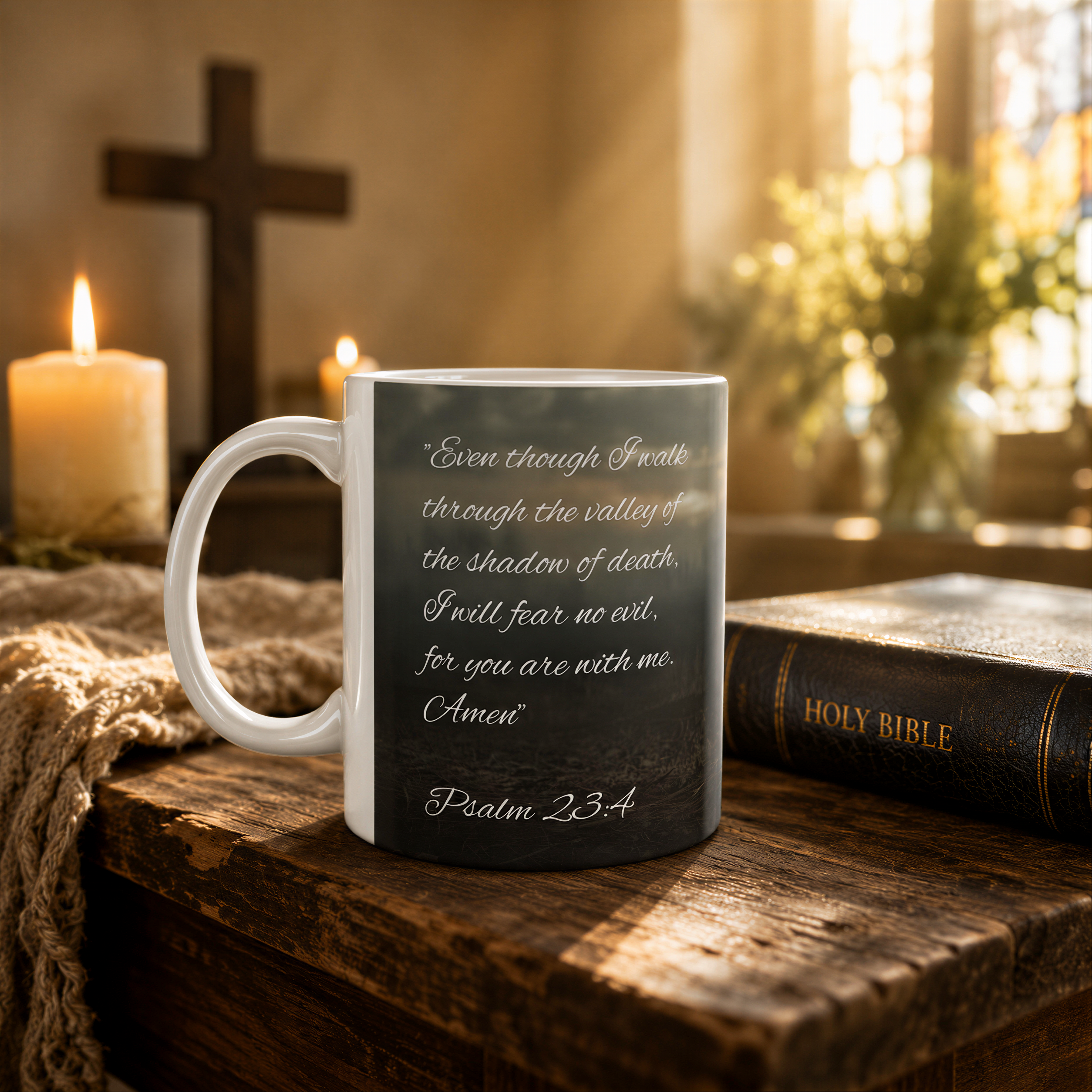 A white mug with a black and white Bible verse printed on it, placed on a wooden surface with a lit candle and a vase of flowers in the background.
