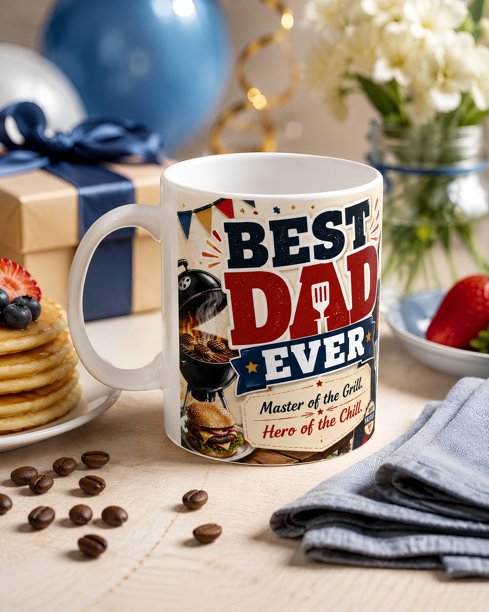 A white mug with a design that says "BEST DAD EVER" and "Master of the Grill" is placed on a table surrounded by pancakes, coffee beans, and a vase of flowers.