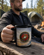 A man is sitting outdoors, holding a black mug with a design featuring the United States Marine Corps emblem.