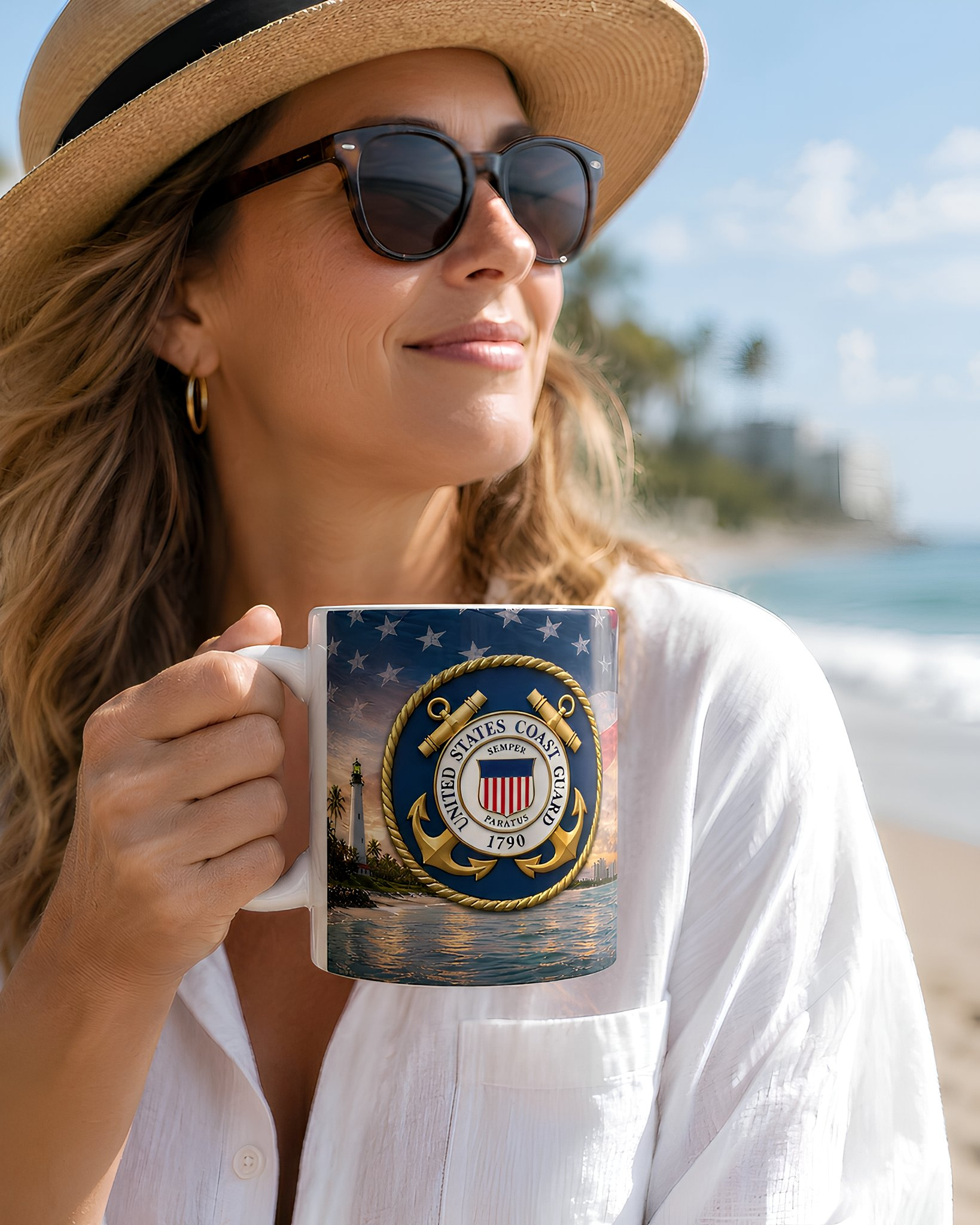 A woman wearing a straw hat and sunglasses holds a mug with a navy blue and gold design featuring a lighthouse and the words "United States Coast Guard" on a beach.