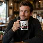 A man is sitting at a table in a coffee shop, holding a mug with a cartoon character on it.