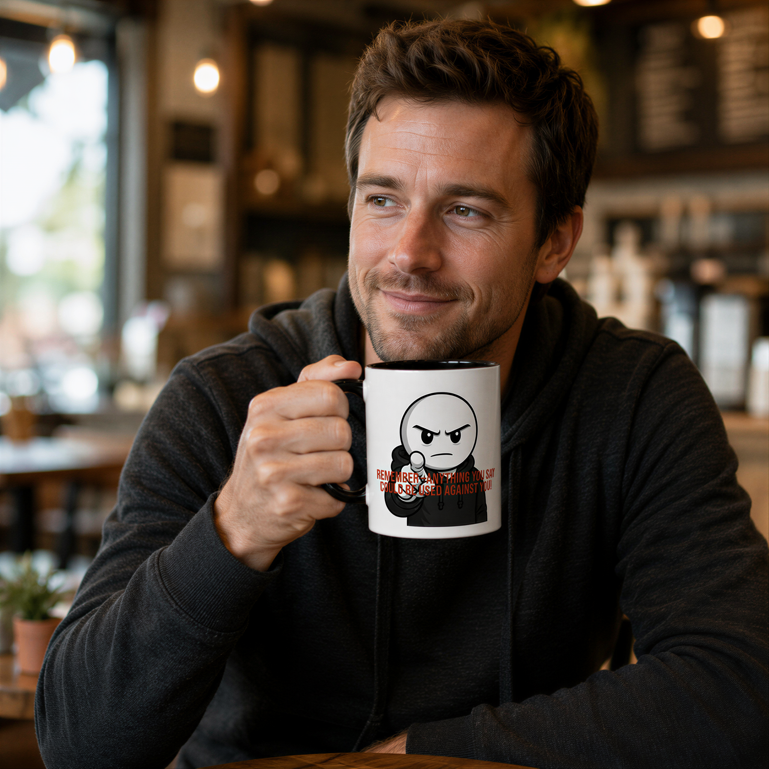 A man is sitting at a table in a coffee shop, holding a mug with a cartoon character on it.